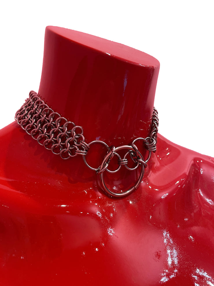 A close-up of a chain mail choker featuring intricate metal links, with bold O-rings positioned at the center. The choker rests elegantly on a neutral backdrop, highlighting its edgy, industrial design.