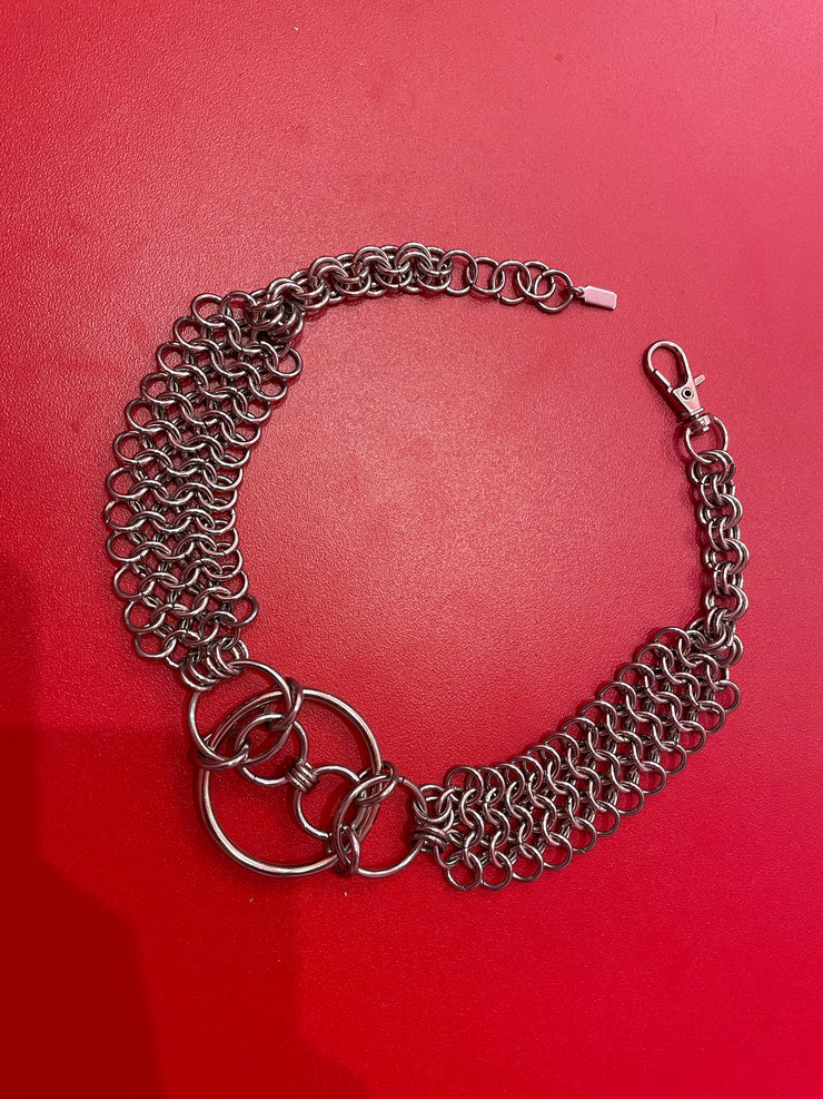 A close-up of a chain mail choker featuring intricate metal links, with bold O-rings positioned at the center. The choker rests elegantly on a neutral backdrop, highlighting its edgy, industrial design.