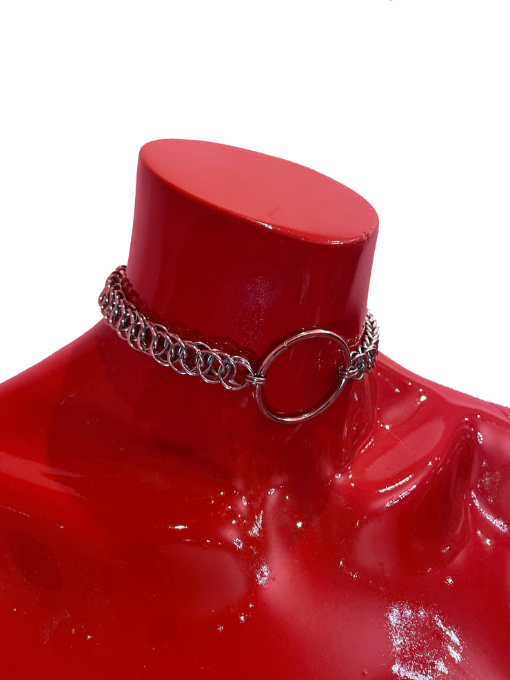 A close-up of a simple silver chain choker with a bold ring at the center. The sleek, minimalist design rests elegantly against a neutral backdrop, highlighting its refined and timeless appeal.