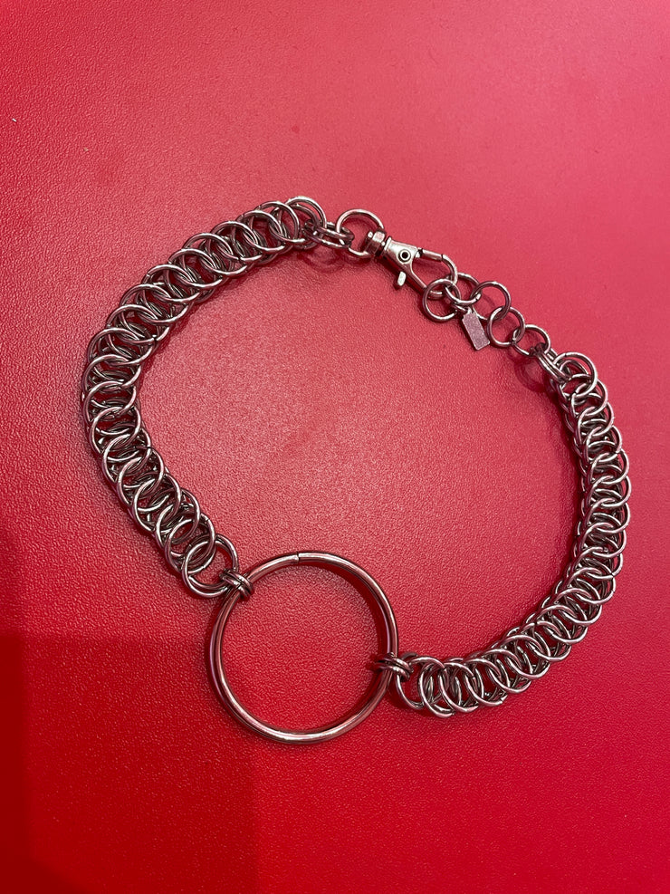 A close-up of a simple silver chain choker with a bold ring at the center. The sleek, minimalist design rests elegantly against a neutral backdrop, highlighting its refined and timeless appeal.