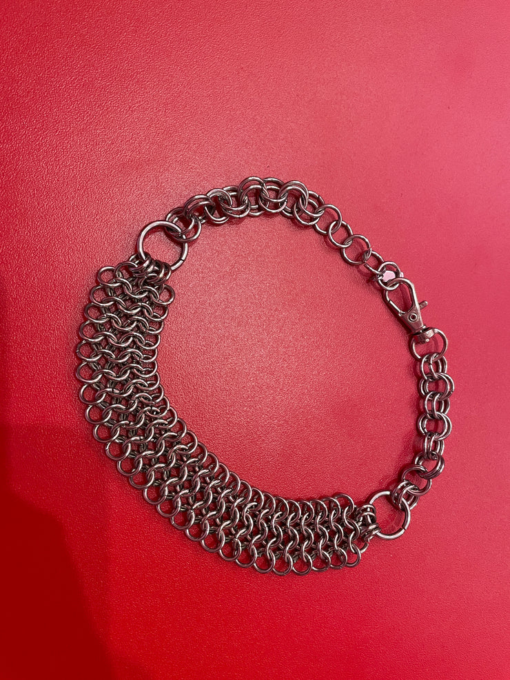 A close-up of a chunky yet lightweight silver chain mail choker, featuring intricate links. The choker rests against a neutral background, showcasing its bold design and adjustable clasp.
