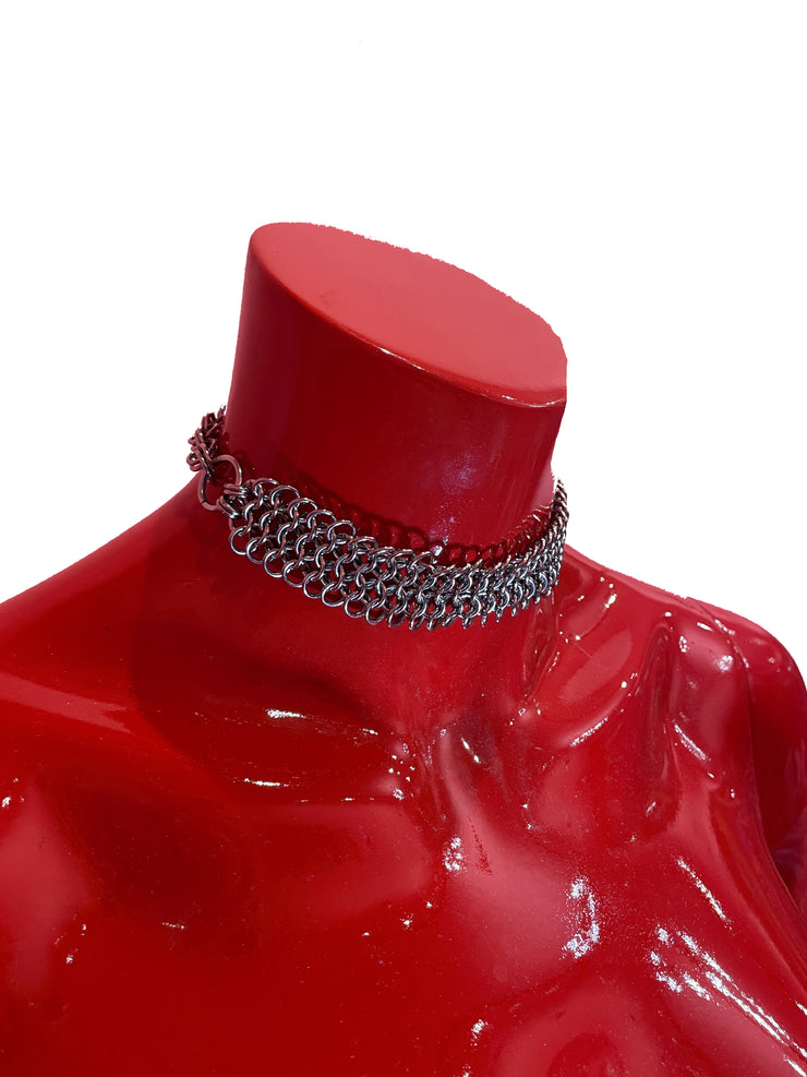 A close-up of a chunky yet lightweight silver chain mail choker, featuring intricate links. The choker rests against a neutral background, showcasing its bold design and adjustable clasp.